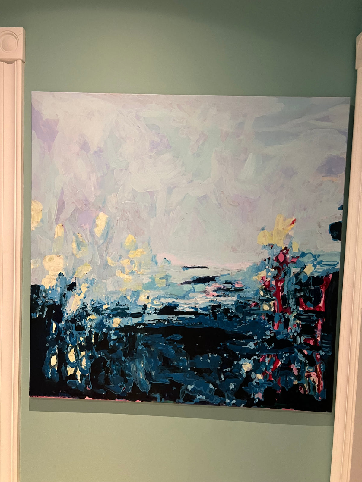 An abstract landscape painting on canvas, featuring a pale lavender and white sky with dark blue, black, and yellow brushstrokes below, displayed on a sage green wall.